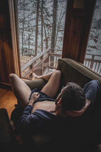 A vertical shot of nude art featuring a Caucasian male laying down on a couch looking out the window