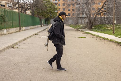 A young caucasian male from Italy with a black coat and cap walking on the street with a skateboard