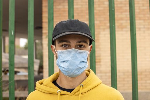A portrait of a young caucasian male from Italy wearing a medical mask standing in front of a green fence
