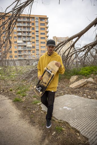 a young caucasian male from Italy in a yellow hoodie and cap holding a skateboard in his hands in a street