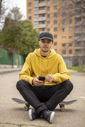 A young caucasian male from Italy in a yellow hoodie and black cap sitting on a skateboard on the street