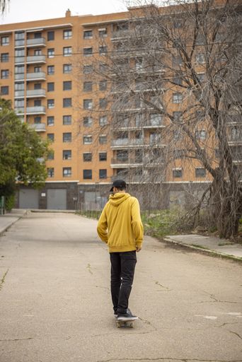 A male in a yellow hoodie and black cap on the skateboard in the background of a high rise building
