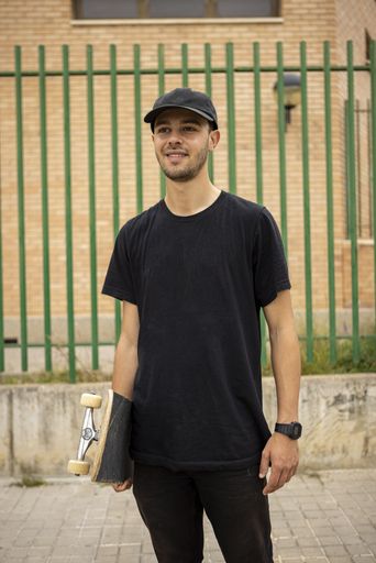A young caucasian male from Italy in a black shirt and cap holding a skateboard in his hand on the stree