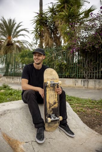 A young caucasian male from Italy with a smile sitting on the street holding a skateboard