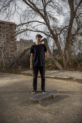 A young caucasian male from Italy in a black shirt and cap standing by his skateboard