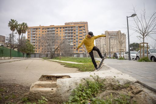 A young caucasian male from Italy in a yellow hoodie and black cap jumping with his skateboard