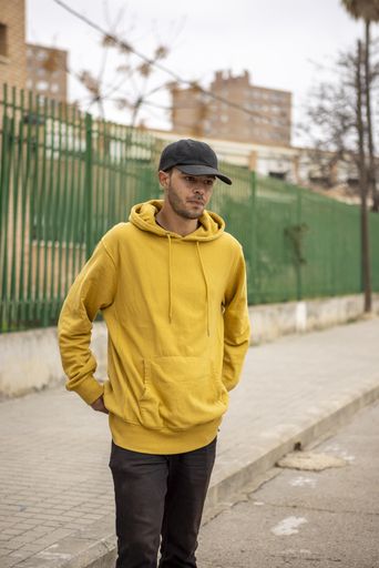 A young caucasian male from Italy in a yellow hoodie and black cap standing on the street