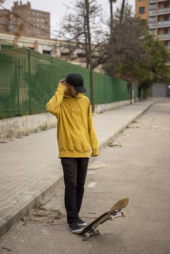 A young caucasian male from Italy in a yellow hoodie and black cap standing on his skateboard