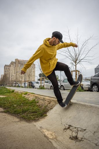 A young caucasian male from Italy in a yellow hoodie and black cap jumping with his skateboard