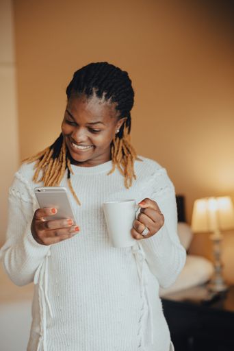 A young African female joyfully using her smartphone while holding a cup of coffee in her hands