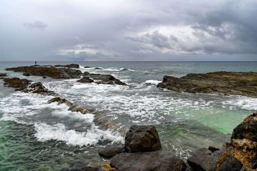 A person standing on the rocks in the sea and fishing on the coast under the cloudy stormy sky