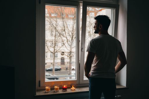 A man standing by the window of his home, looking outside, as the warm flicker of lit candles illuminates the interior