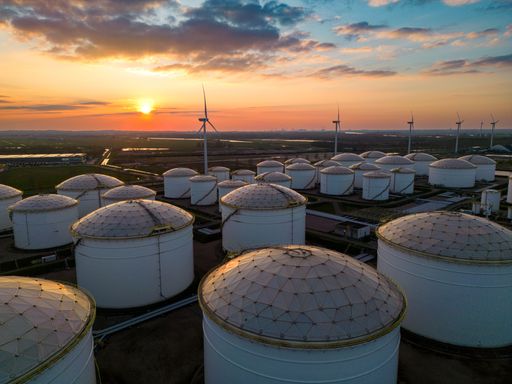 An aerial shot of the sunset behind an oil field filled with tanks and wind turbines.