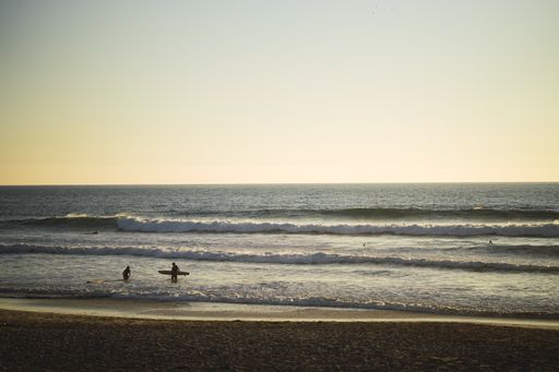 A scenic view of a surfer walking on a beach carrying a surfboard under a clear sky
