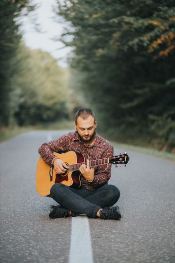 A vertical shot of a White guy playing the guitar sitting on a road in Brcko, Bosnia and Herzegovina