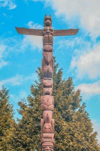 A vertical shot of totem pole surrounded with greenery under cloudy blue sky