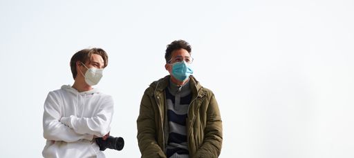 A panoramic shot of two young Caucasian and Hispanic guys wearing sanitary masks outdoors