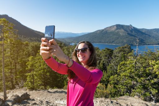 A Caucasian woman taking a selfie of a mountain lake on her smartphone