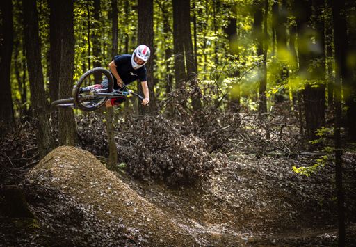 A man in his mid-twenties is performing a jump on a mountain bike while riding on a dirt trail in a wooded area