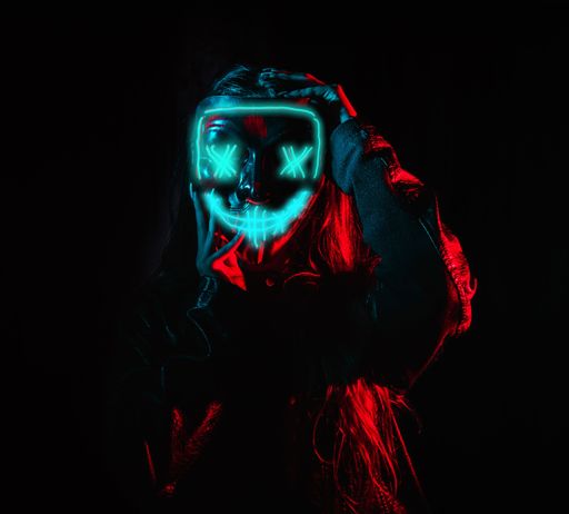 A crazy girl with her fingers on her ominous neon mask striking a pose in a dark room