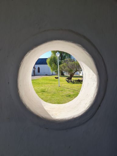 A round opening in an exterior wall providing a view of a nearby church with a blue sky backdrop