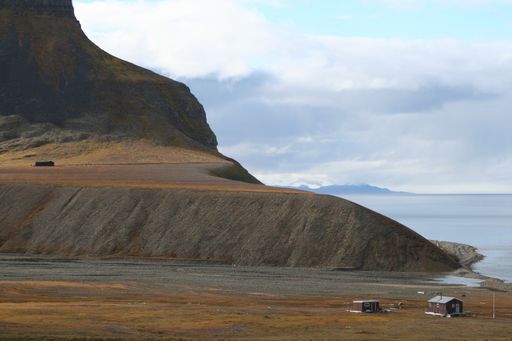 Two small houses on a grassy field near a large rock formation overlooking a body of water
