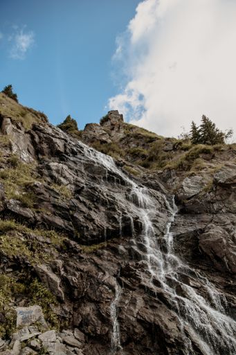 A waterfall surrounded by rocks covered in greenery under the sunlight and a blue cloudy sky