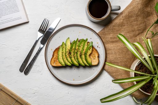 A top view of a delicious avocado toast on a white plate, a cup of coffee, and a book next to it