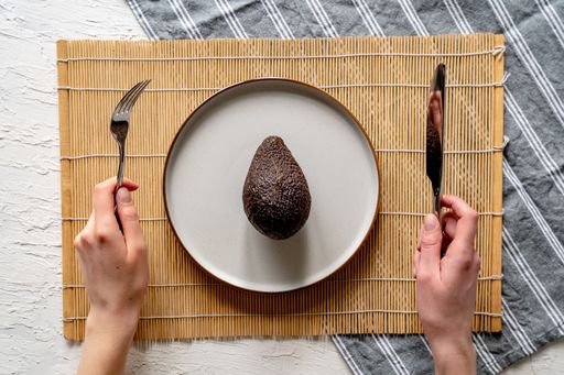 A top view of fresh whole avocado on a white plate with someone holding a fork and knife