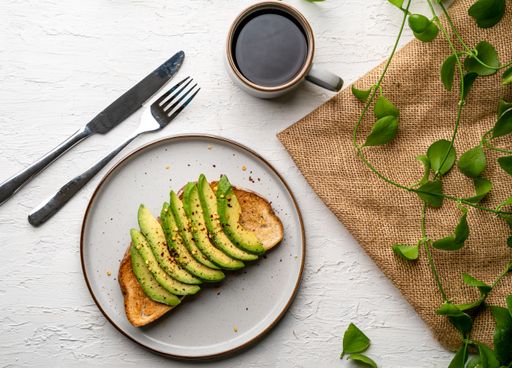 A top view of a delicious avocado toast on a white plate, a cup of coffee next to it