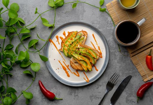 A top view of a delicious avocado toast covered in spicy sauce, a cup of coffee, and plants
