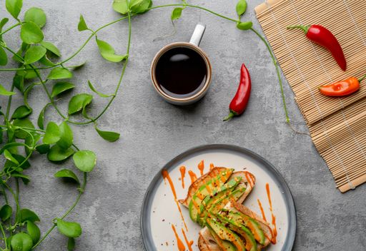 A top view of a delicious avocado toast covered in spicy sauce, a cup of coffee, and plants