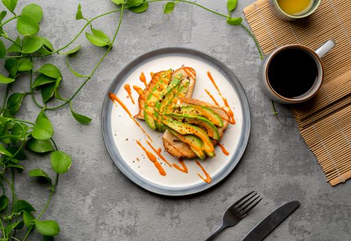 A top view of a delicious avocado toast covered in spicy sauce, a cup of coffee, and plants