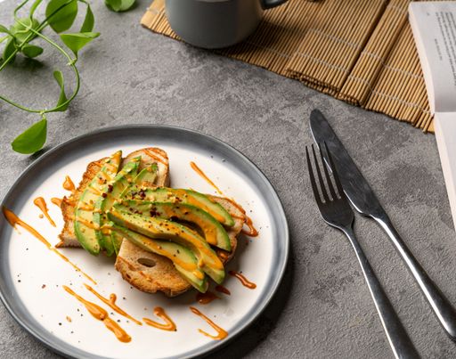 A closeup of a delicious avocado toast covered in spicy sauce on a white plate, with fork and knife
