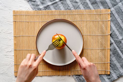 A top view of a fresh apple on a white plate, with someone using the fork and knife