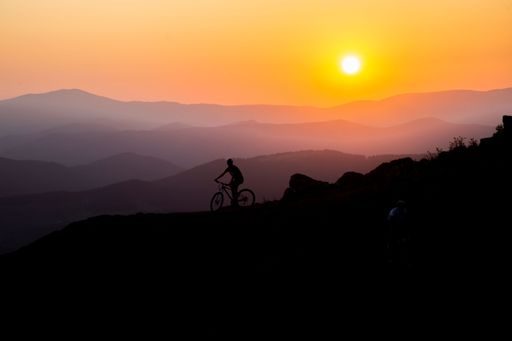 A silhouette of a person riding a bike up a hill at sunset with majestic mountains in the backdrop.