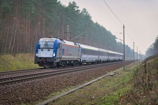 A vintage train moves along the tracks in a landscape, with lush green trees in the background