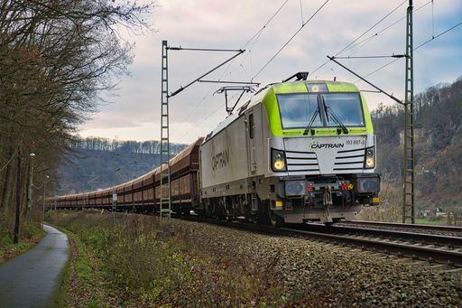 A train moves along the tracks in a landscape, with lush green trees in the background