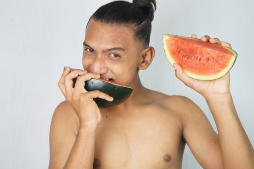 A portrait of a topless man from India with a man bun biting the slice of watermelon in his hands while looking at the camera