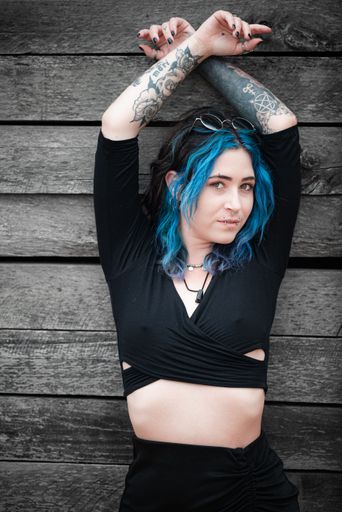 A young Caucasian female with blue hair standing against a wooden backdrop