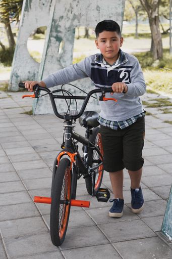 A front view of a hispanic smiling cute  child standing at the park bnext to his bicycle looking at the camera