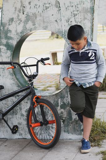 A front view of a hispanic smiling cute child leaning against the wall at the park next to his bicycle