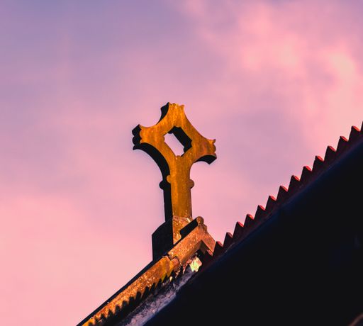 An isolated image of a stone cross on top of a roof, with a pink sky in the background