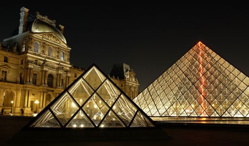 Two of the famous Louvre Pyramids at night, lit by golden lights, with the main building of the Museum in the background.