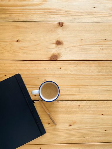 A closeup of a closed notebook, a pen, and a cup of coffee on a wooden table with free space for text