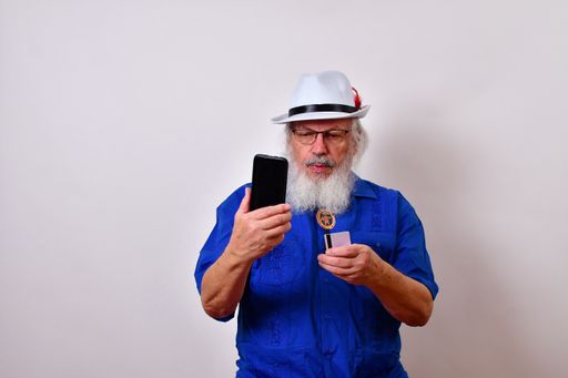 A mature gentleman with a white fedora hat, blue guayabera shirt, and bolo tie doing online commerce using his cellphone and a debit card
