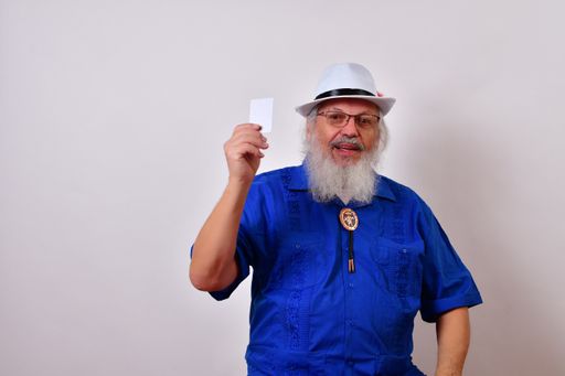 A mature gentleman with a white fedora hat, blue guayabera shirt, and bolo tie holding up his credit card isolated on a white background
