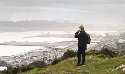 An adult male backpacker standing on a hill and talking on the phone while looking at the sea in Spain