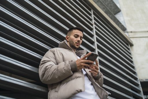 A low angle shot of a sexy male in a street style outfit using his phone outdoors