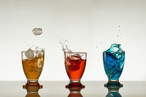 the ice cubes falling into a glass of yellow drink next to glasses of red and blue drinks